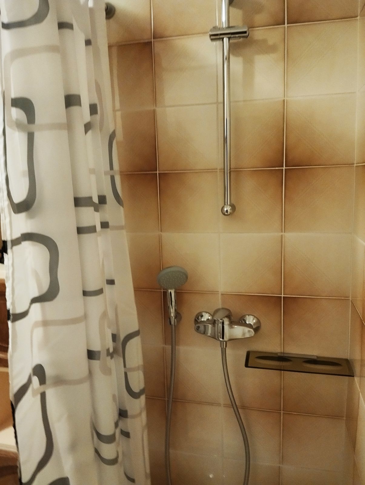 A bathroom with beige tiled walls, a shower with a handheld showerhead, and a curtain with a geometric pattern.