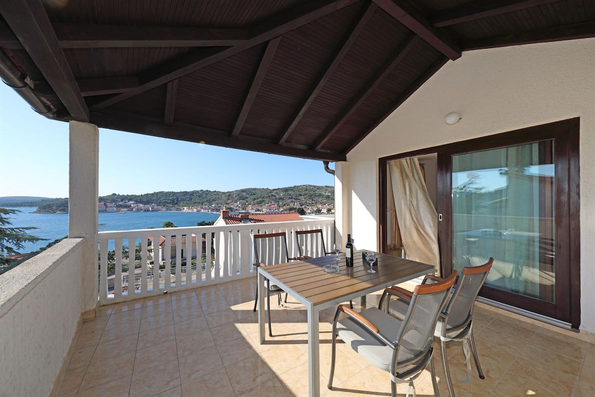 A balcony with a table and chairs, overlooking a scenic view of the sea and hills.