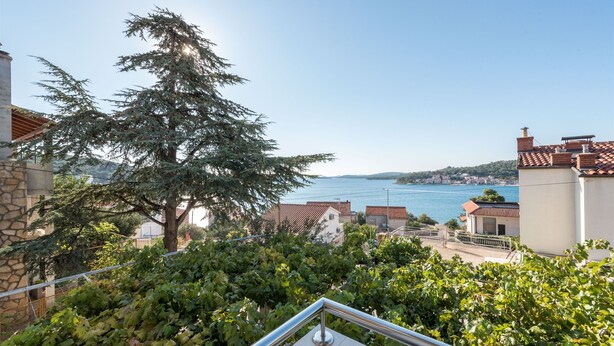 Un balcon doté d’une balustrade en métal, donnant sur une ville côtière, avec un grand arbre à feuilles persistantes et une vue sur la mer.