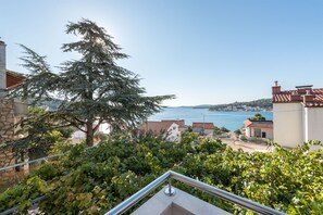 Un balcón con barandilla metálica que da a un pueblo costero, un árbol de hoja perenne y vistas al mar.