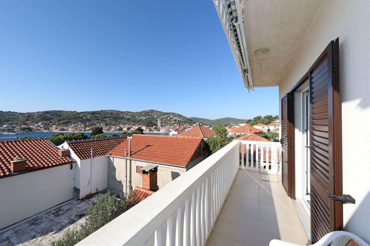 A balcony with a view of a coastal town, white railing, and a door with shutters.