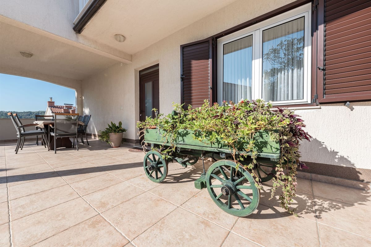 A patio with a table and chairs, a green cart with plants, and a view of the sea.