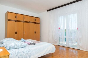Quarto com cama, guarda-roupa de madeira e uma janela grande com cortinas leves.