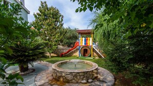 Une fontaine en pierre, entourée d’un chemin également en pierre, d’une aire de jeux colorée et d’une végétation luxuriante.