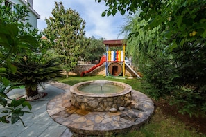 Une fontaine en pierre entourée d’une terrasse en pierre, un terrain de jeux avec un toboggan et une végétation luxuriante.