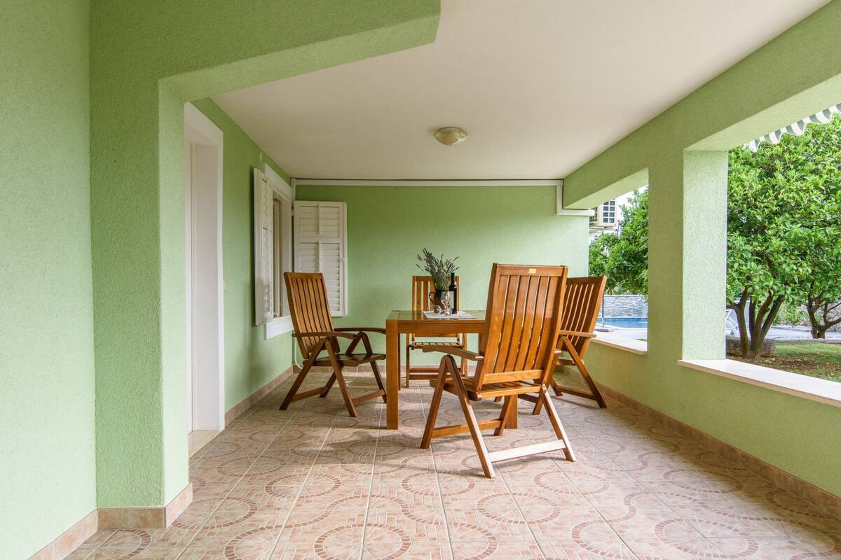 Ein überdachter Patio-Bereich mit einem Tisch und vier Stühlen, ein Fenster mit Blick auf Bäume und eine Tür mit weißen Fensterläden.