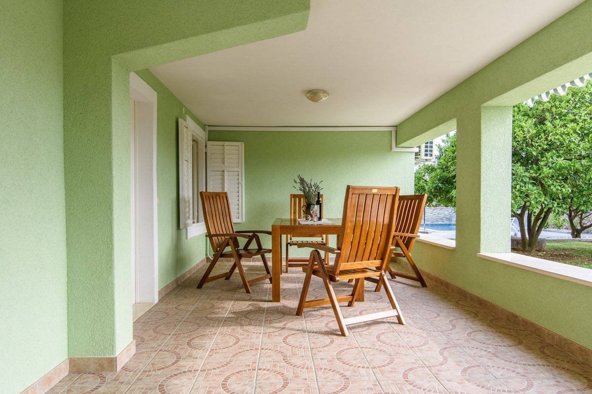 Un patio cubierto con una mesa y sillas, una ventana que da a zonas verdes y una puerta con contraventanas blancas.