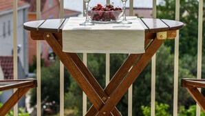 Une table en bois avec deux verres de vin blanc et un saladier de fruits, le tout placé sur un balcon offrant une vue sur les montagnes et les bâtiments.