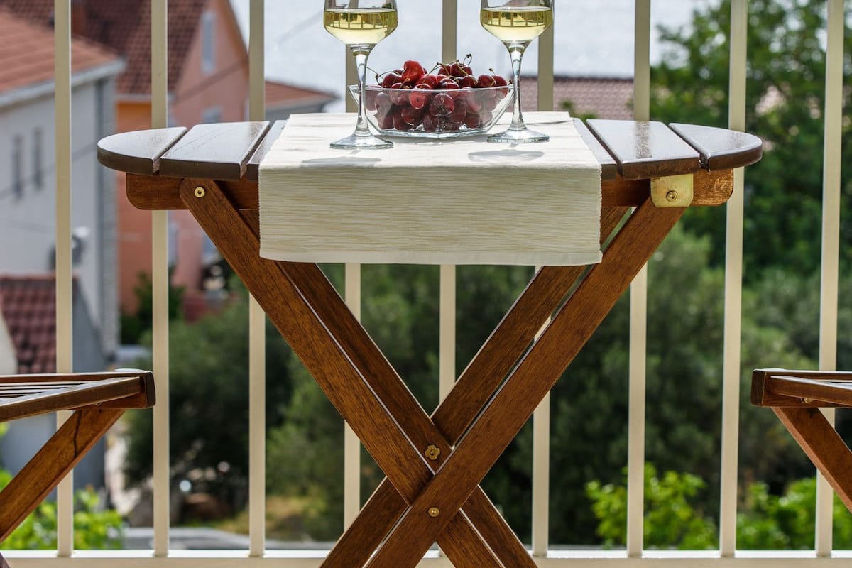 Ein Holztisch mit zwei Weingläsern und einer Obstschale auf einem Balkon mit Blick auf Berge und Gebäude.