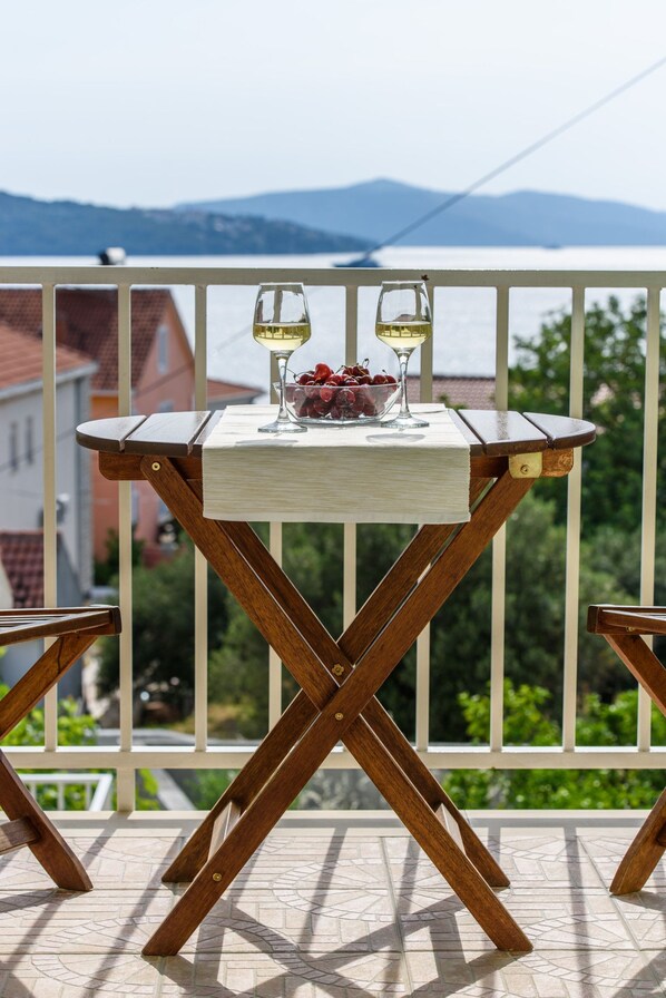 Una mesa de madera con dos copas de vino blanco y un cuenco de fruta en un balcón con vistas a montañas y edificios.