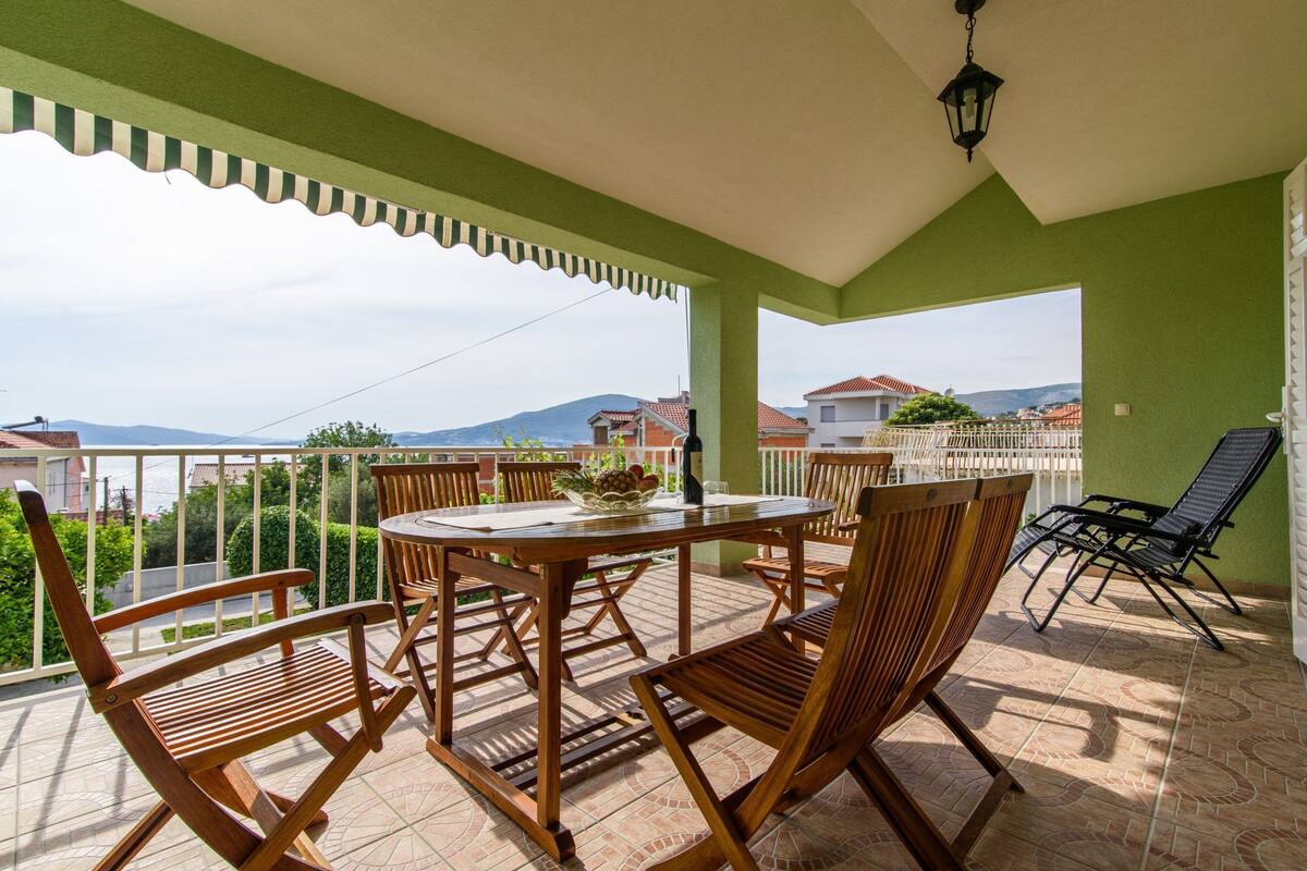 Eine überdachte Terrasse im Freien mit einem runden Tisch und Holzstühlen, einem schwarzen Liegestuhl und Blick auf Häuser und Berge im Hintergrund.