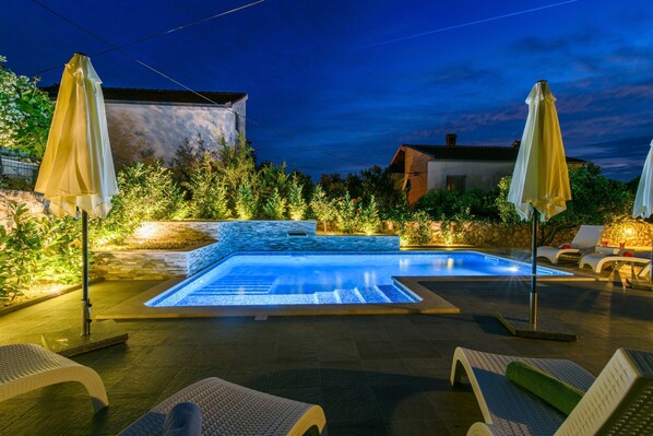 Une piscine entourée de verdure et de murs en pierre, avec des chaises longues et des parasols.