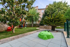 Un espace de jeux pour enfants avec une structure ludique verte en forme de grenouille, un arbre et un bâtiment en arrière-plan.