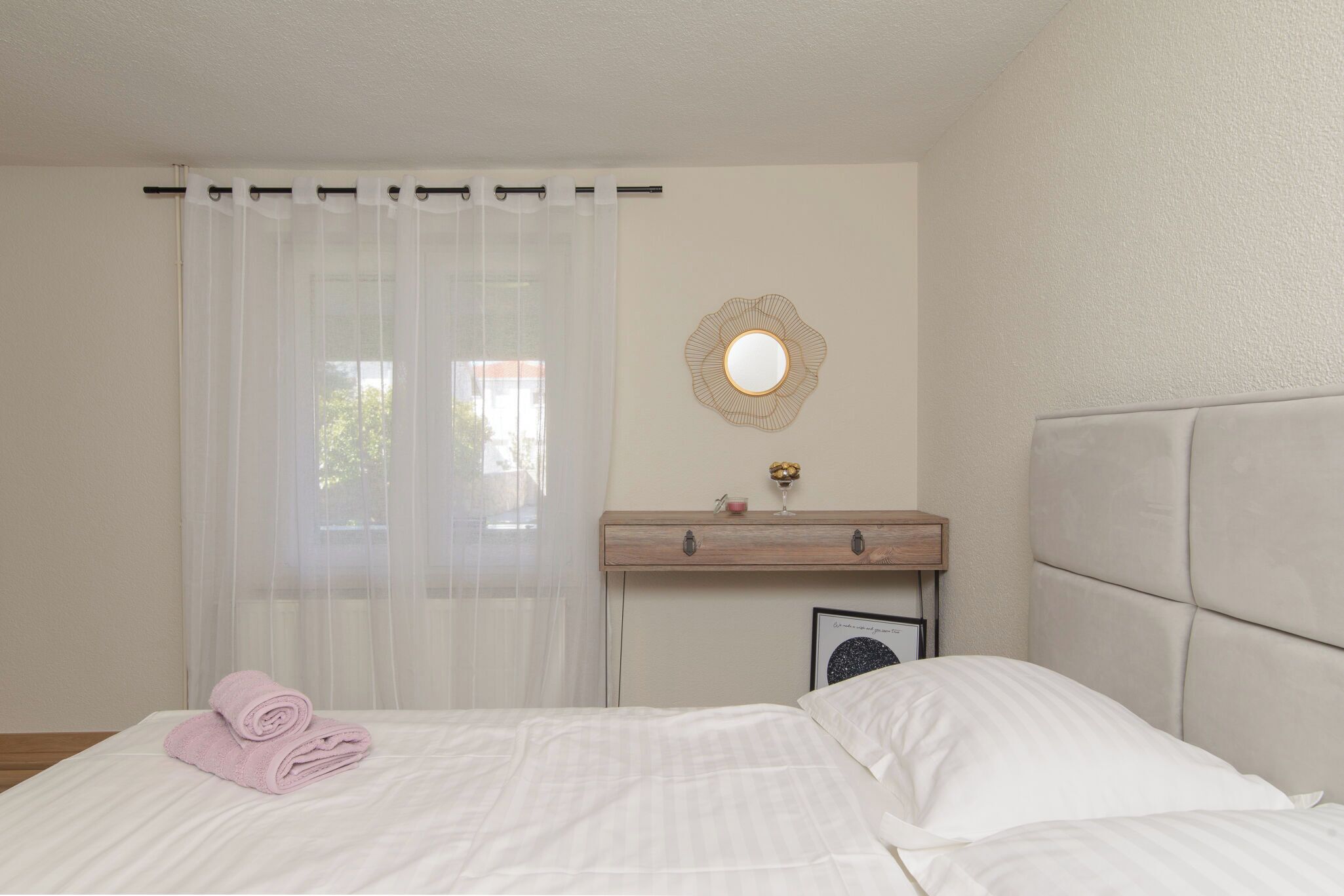 A neatly made bed with white linens, a wooden bedside table with a small decorative item, a wall-mounted mirror, and sheer white curtains.