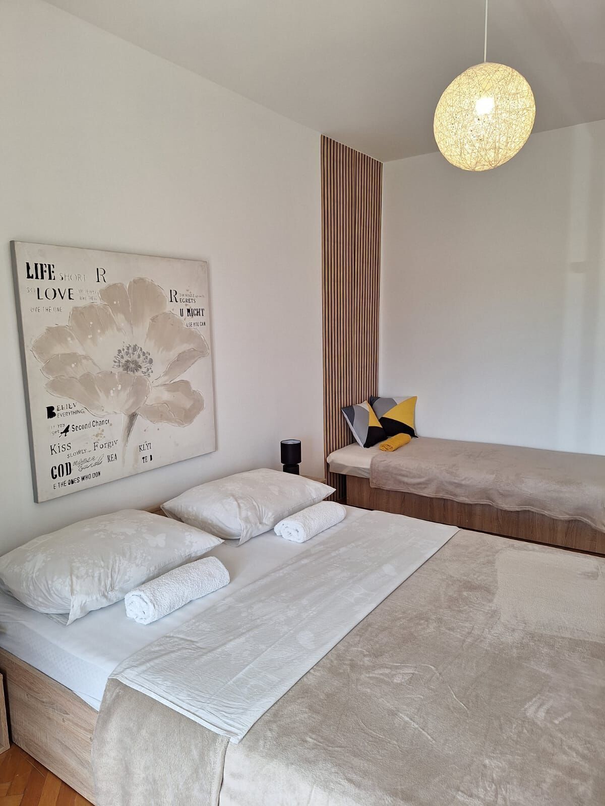 A modern bedroom with two beds, a large wall art piece, a hanging light fixture, and a vertical striped panel.
