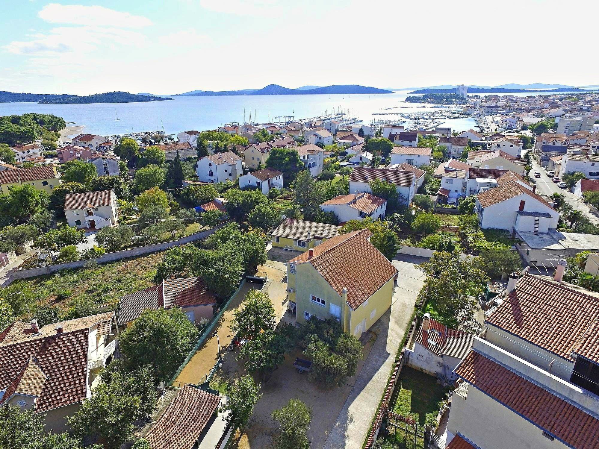 Il s’agit d’une ville côtière où se côtoient des immeubles d’habitation, certains avec des toits recouverts de tuiles rouges et d’autres avec des murs blancs. On aperçoit une route et une étendue d’eau en arrière-plan.