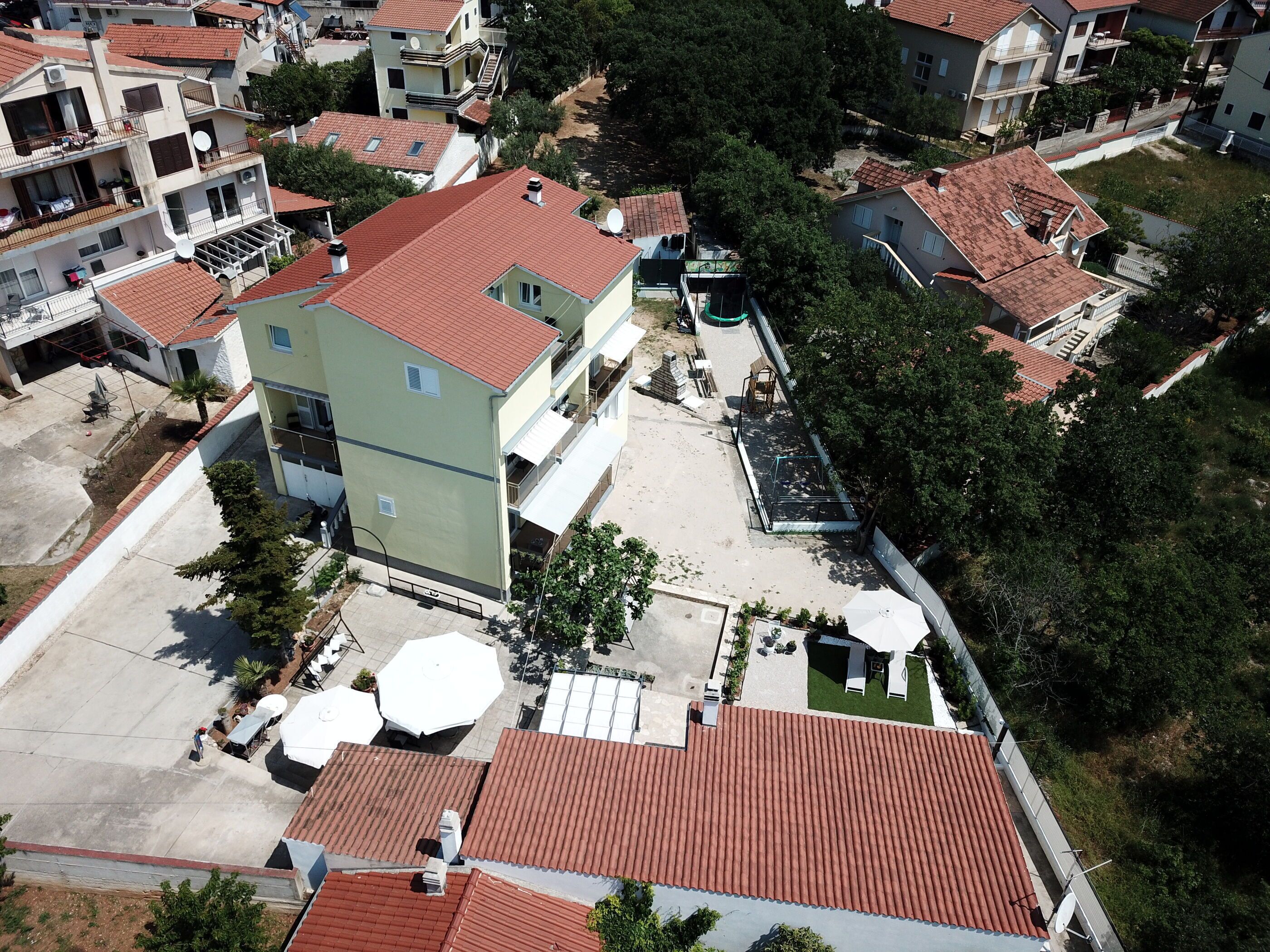 Vista aérea de una zona residencial con varios edificios, incluyendo una casa de dos plantas con techo de tejas rojas y un edificio de techos planos con balcones.
