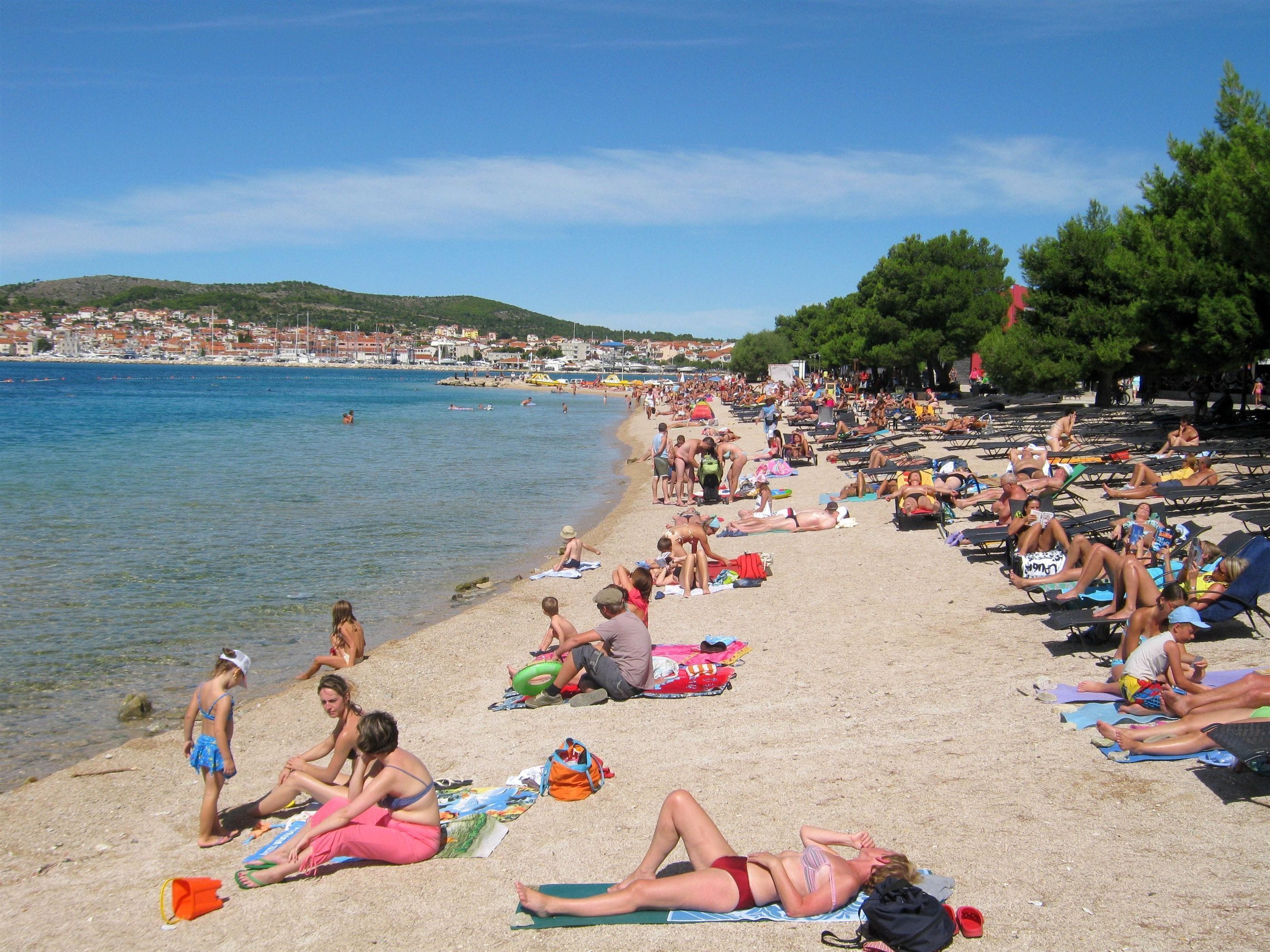 A crowded beach with people sunbathing and swimming, a clear sky, and a distant cityscape.