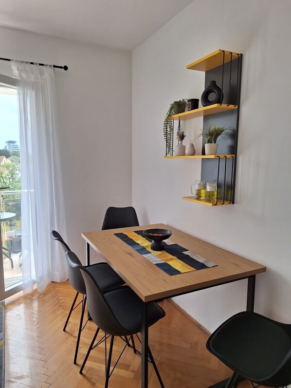 A dining area with a wooden table, black chairs, and a yellow and black patterned runner. There are black shelves with plants and glasses on the wall. - Apartments Ivona (Vodice)
