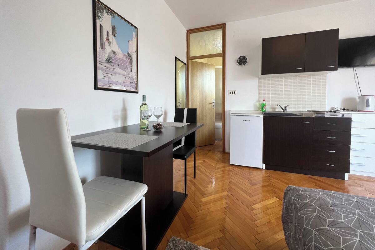 A modern kitchen with a white refrigerator, dark cabinets, and a flat-screen TV. There is a dining area with a table and chairs, and a painting on the wall.