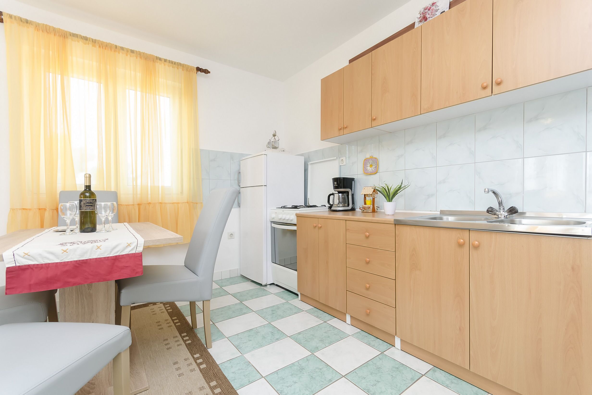 A compact kitchen with wooden cabinets, a white refrigerator, a coffee maker, and a dining table with a red tablecloth.