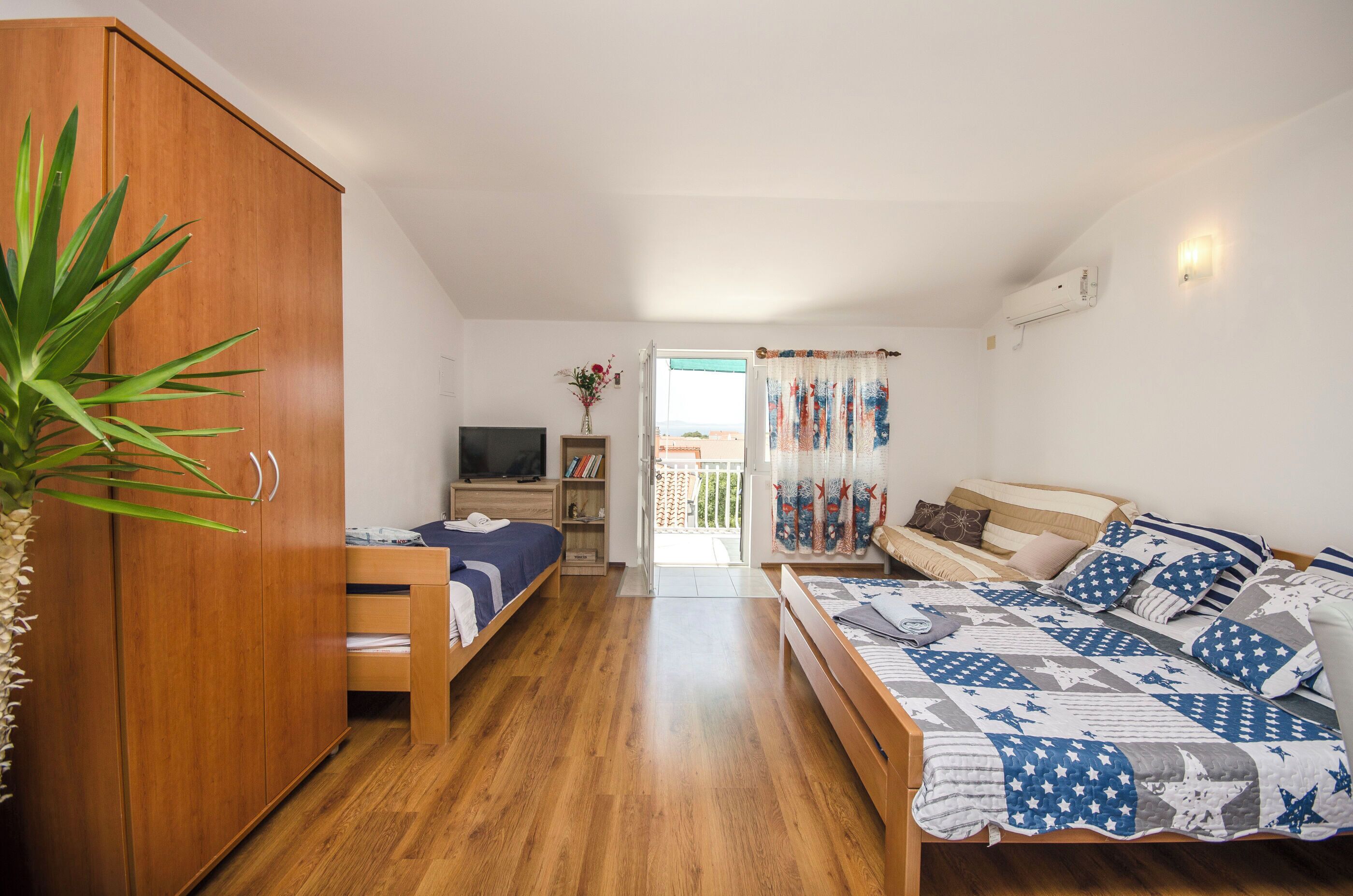 A bedroom with a wooden wardrobe, a bed with a blue and white quilt, a TV, and a plant.