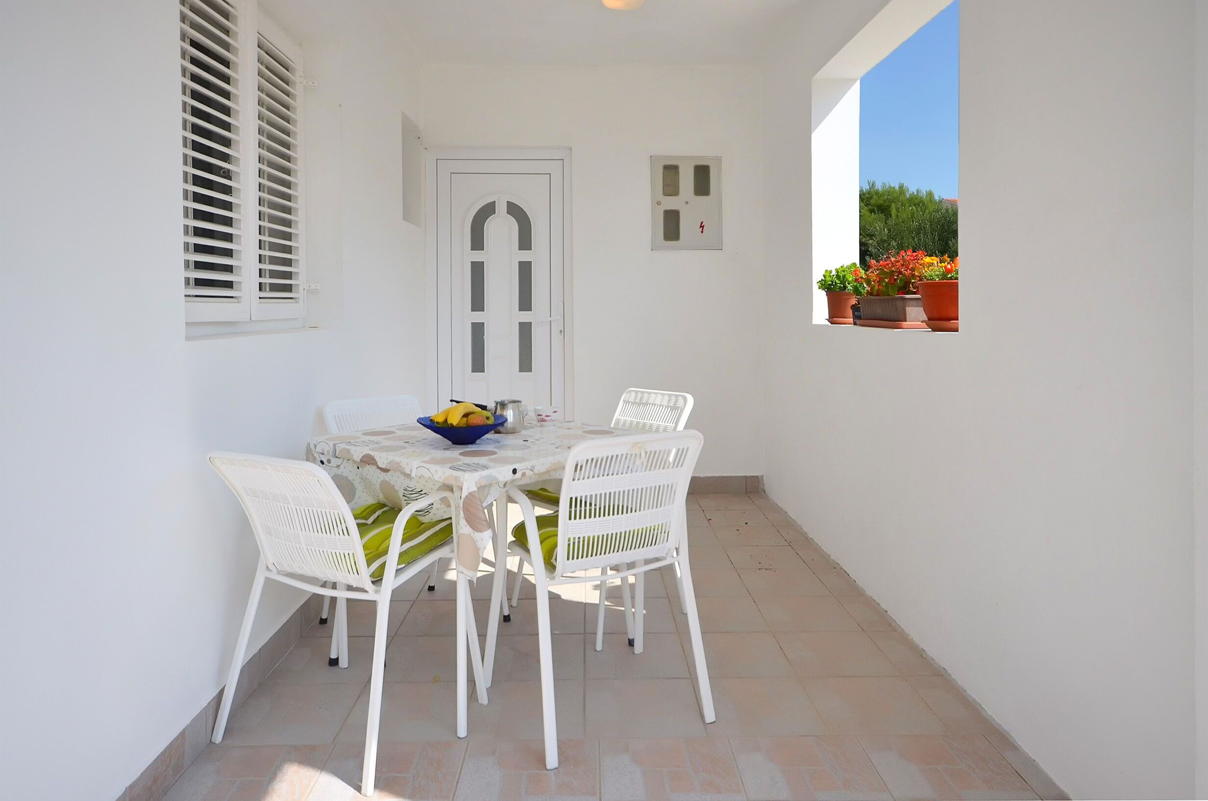A small outdoor patio with a table and chairs, a door, and a window with potted plants.