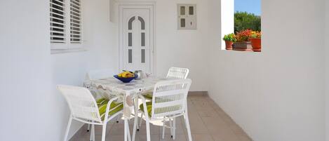 A small outdoor patio with a table and chairs, a door, and a window with potted plants.
