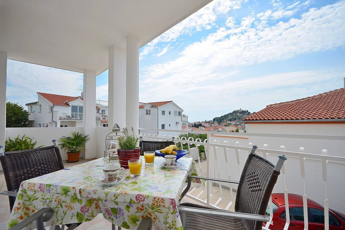 A balcony with a table set for breakfast, potted plants, and a red car parked nearby.
