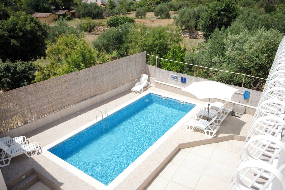 A rectangular swimming pool with a tiled deck, surrounded by white lounge chairs and a white umbrella.