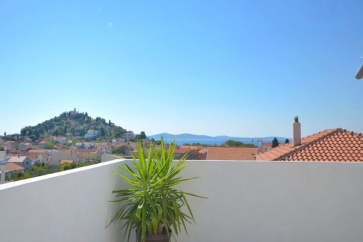A view of a town with a hilltop church, terracotta-roofed buildings, and a potted plant in the foreground.