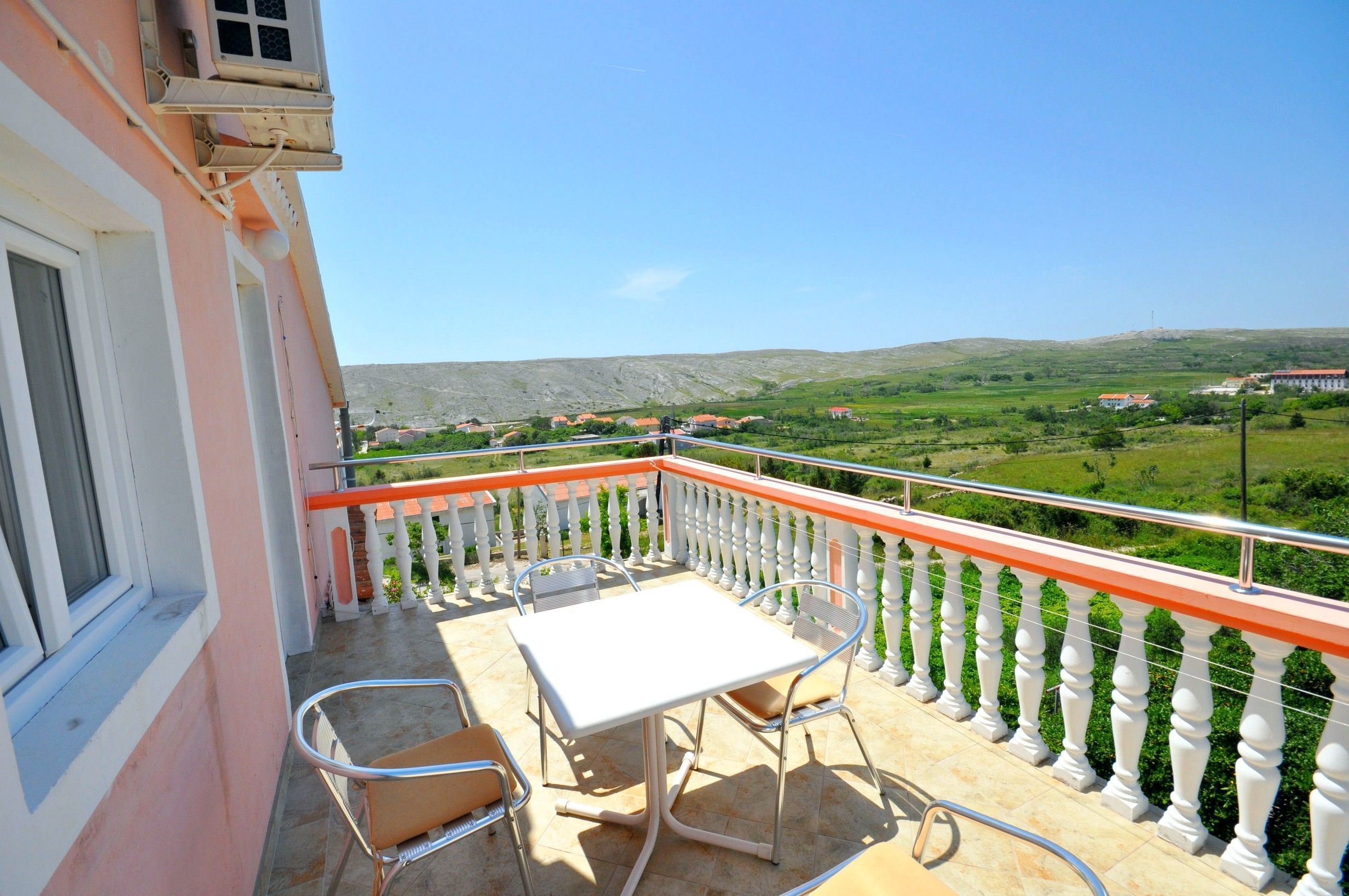 Un balcone con tavolo e sedie, affaccia su un paesaggio verde con edifici in lontananza.