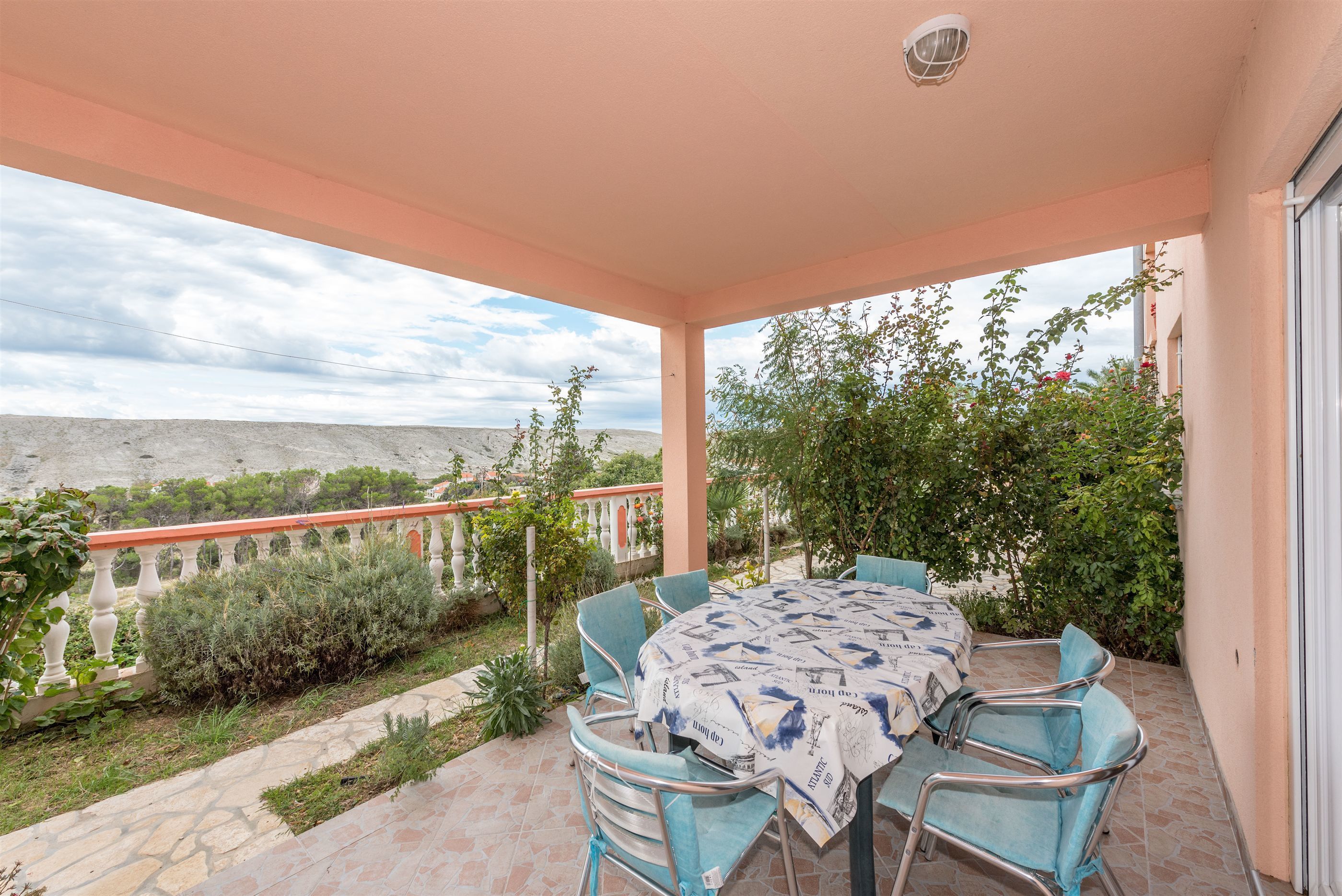 A covered outdoor patio with a table and chairs, a view of the landscape, and a railing.