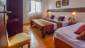 A hotel room with two beds, a wooden chair, a window with sheer curtains, and wall-mounted lamps.