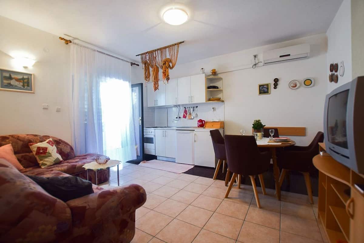 A living room with a sofa, a dining table with chairs, a kitchen area with cabinets, and a television.
