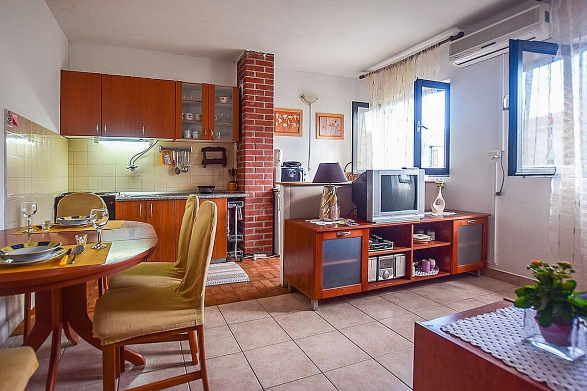 A compact kitchen with wooden cabinets, a dining table with chairs, a television on a wooden stand, and a potted plant.