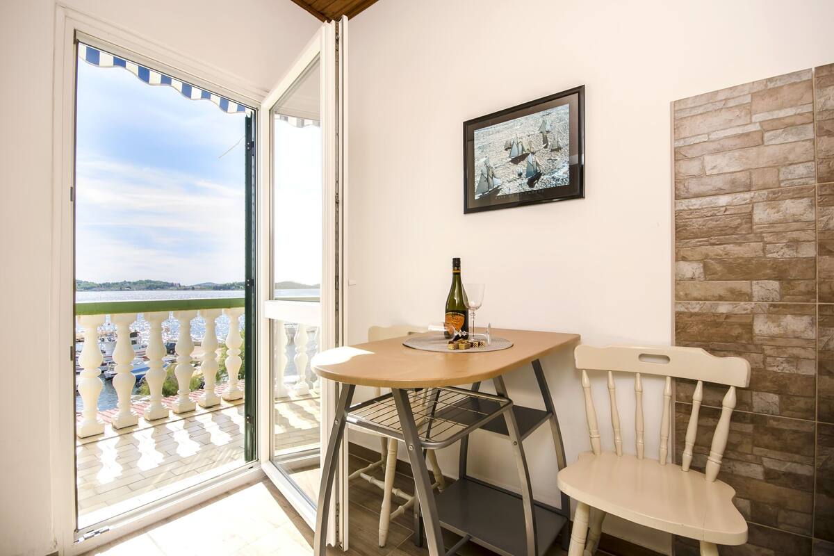 A balcony with a view of a body of water and boats, a table with a bottle and glass, and a chair.