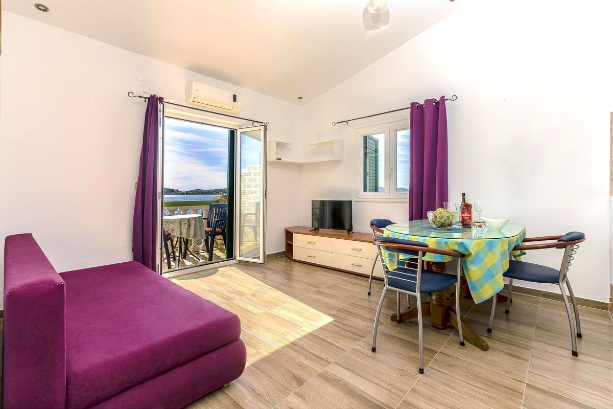 A modern living room with a purple sofa, a dining table with chairs, a TV, and a view of the outdoors through a glass door.