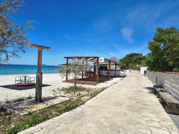 A beachside walkway leading to a wooden structure with a sign, a beach with a pebble surface, and a clear blue sky.