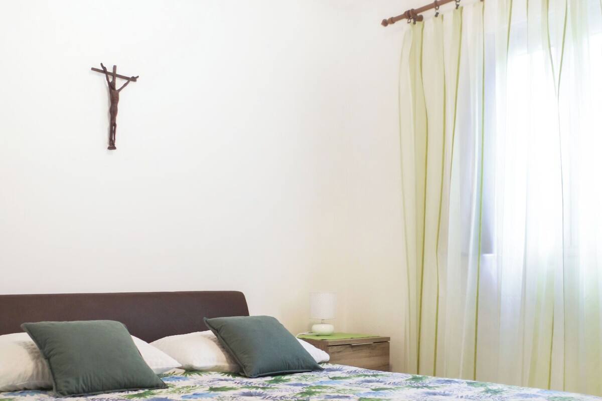 A bedroom with a bed, a nightstand, a wall-mounted cross, and sheer curtains.