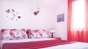 Un dormitorio con una cama, almohadas estampadas, un aplique en la pared, una mesita de noche con lámpara y una ventana con cortinas.