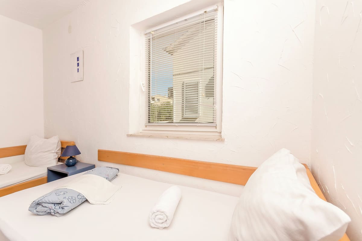 Habitación de hotel con dos camas, una ventana con persianas, una mesita de noche con lámpara y un interruptor de luz en la pared.