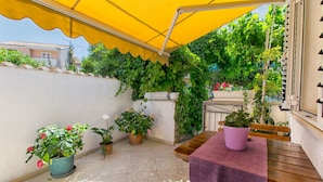 A patio with a yellow awning, potted plants, a wooden table with a purple tablecloth, and a view of a house in the background. - Apartments Kirigin (Supetar)