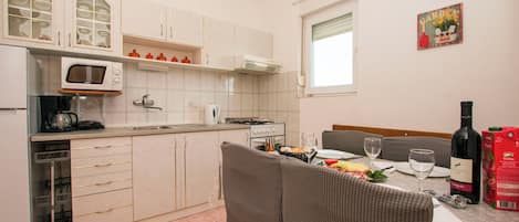 A compact kitchen with a microwave, stove, and sink. There is a dining area with a table set for two, a wine bottle, and a box of cereal.