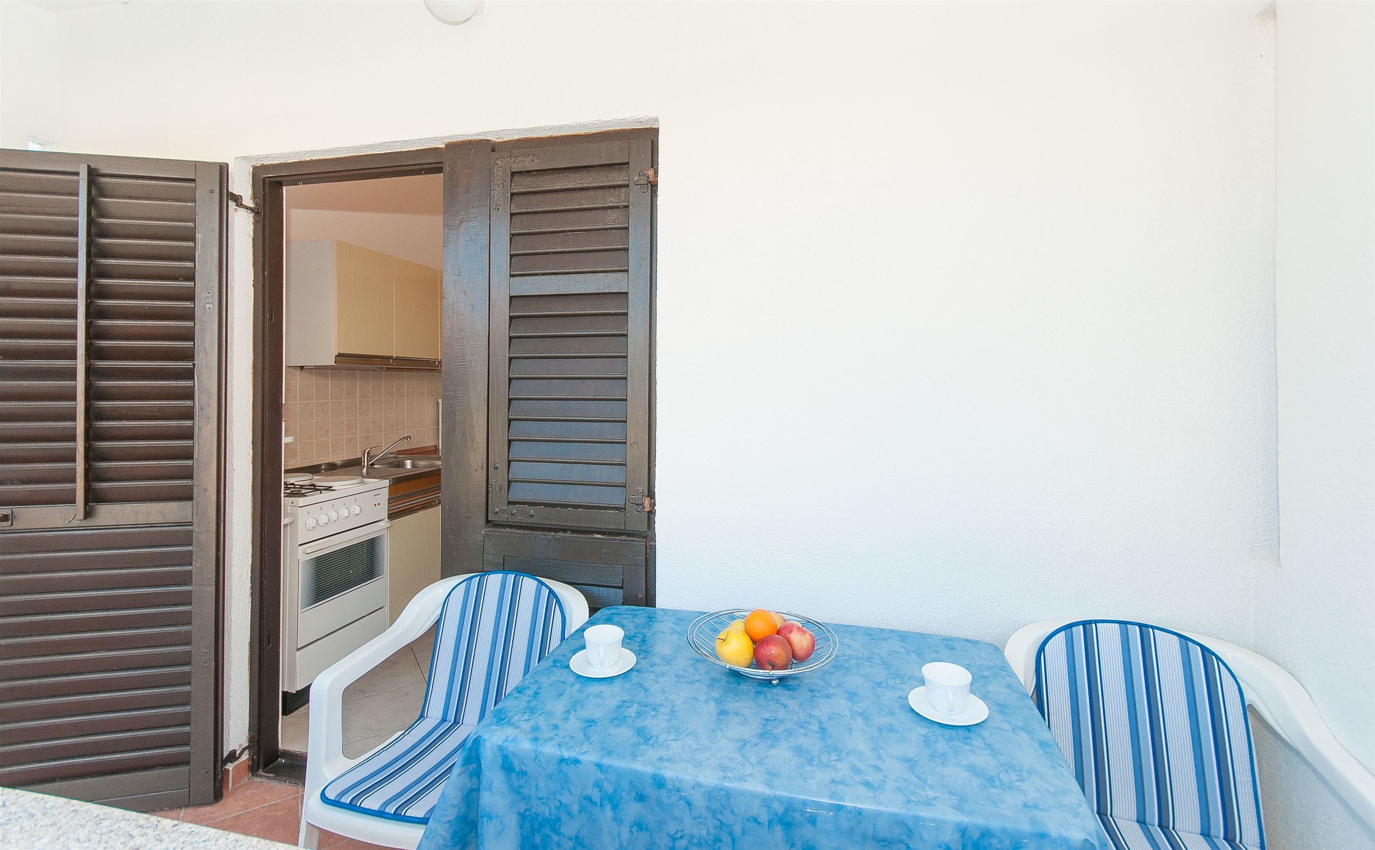 Un pequeño comedor con una mesa lista para dos, un frutero y una cocina visible a través de una puerta abierta.