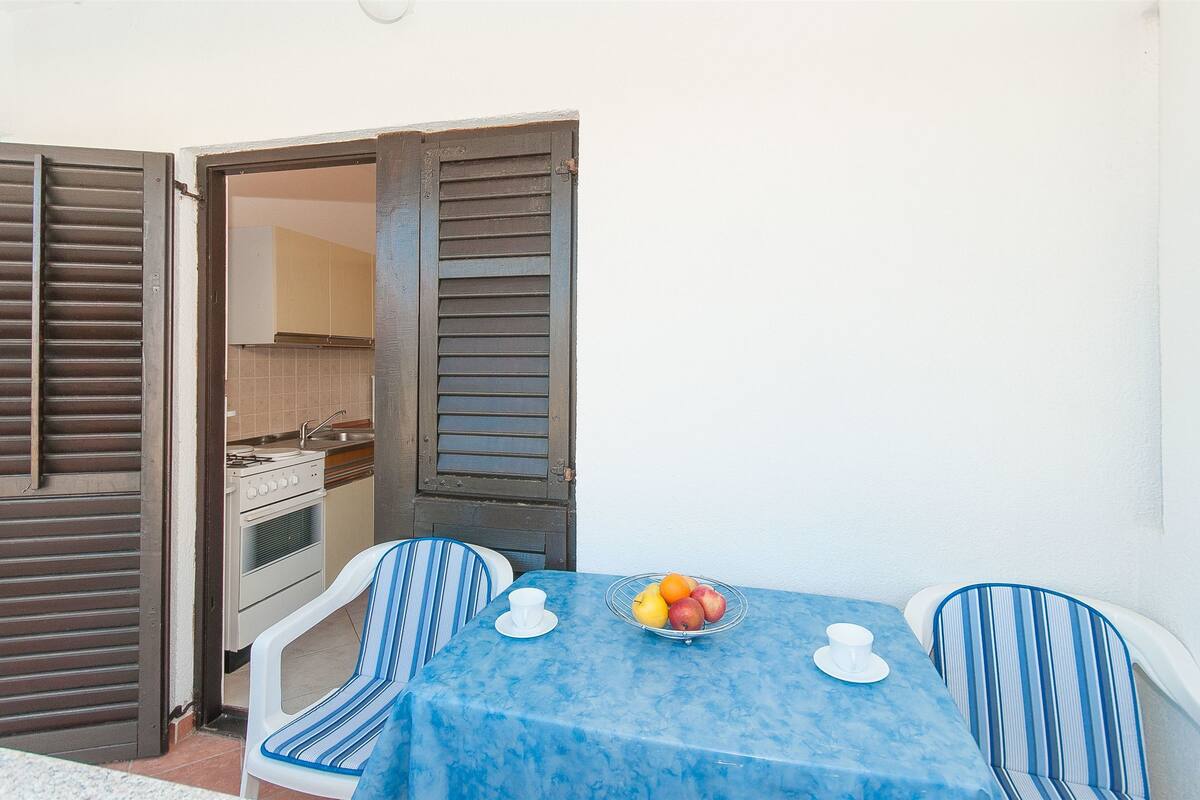 A small dining area with a table set for two, a bowl of fruit, and a kitchen visible through an open door.