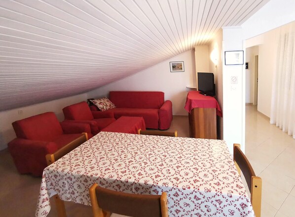 A cozy room with a red sofa, a dining table with a floral tablecloth, and a television on a stand. - Apartments Mate (Pag)
