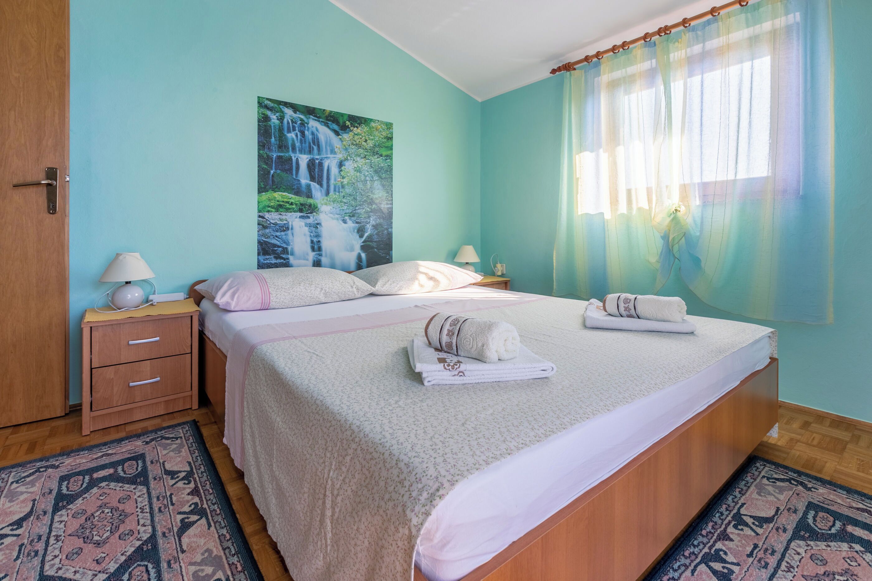 A bedroom with a bed, a nightstand, a lamp, a painting of a waterfall, and a window with curtains.