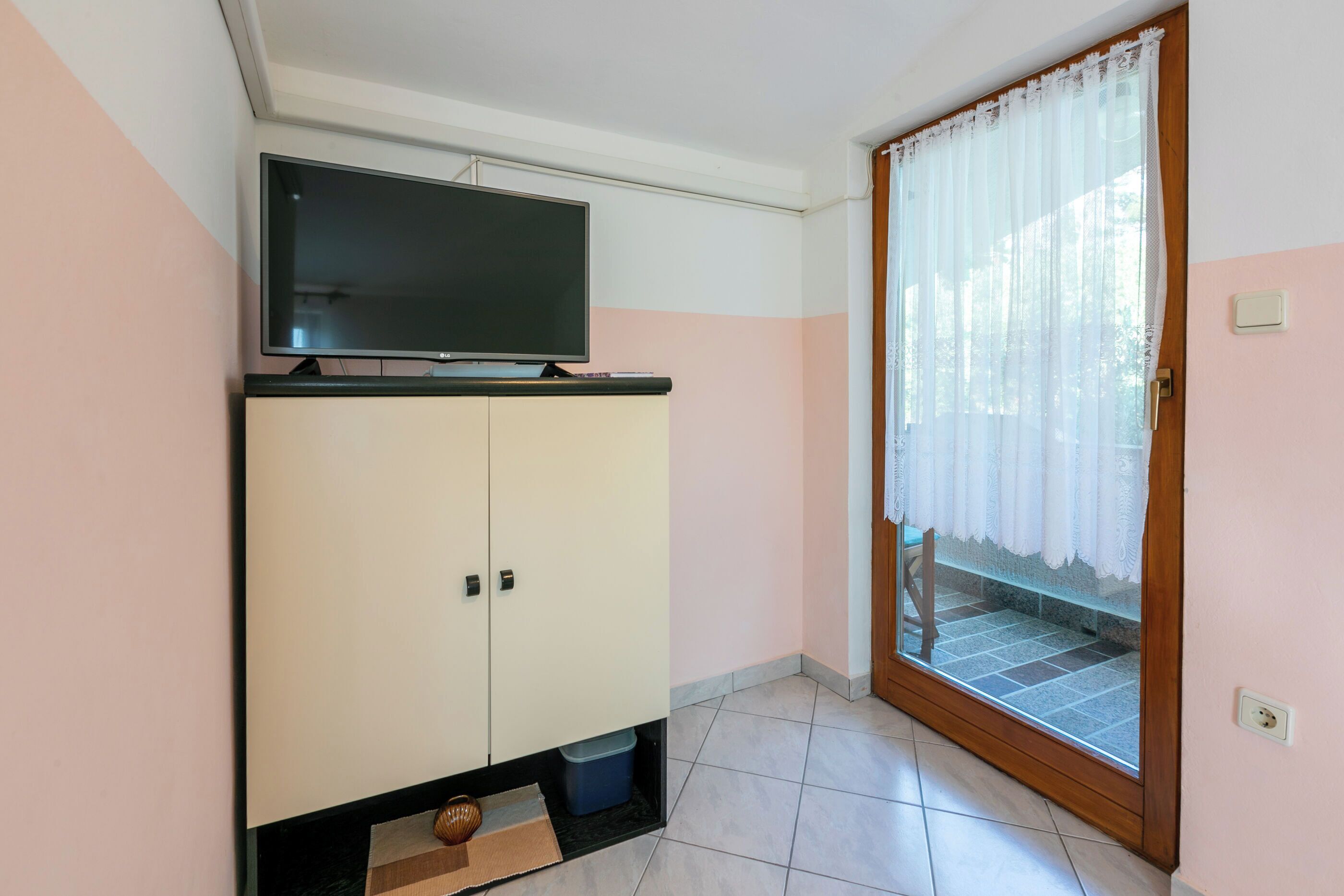 Una habitación con televisión sobre un mueble, una puerta corrediza de vidrio con cortina blanca y suelo de baldosas.