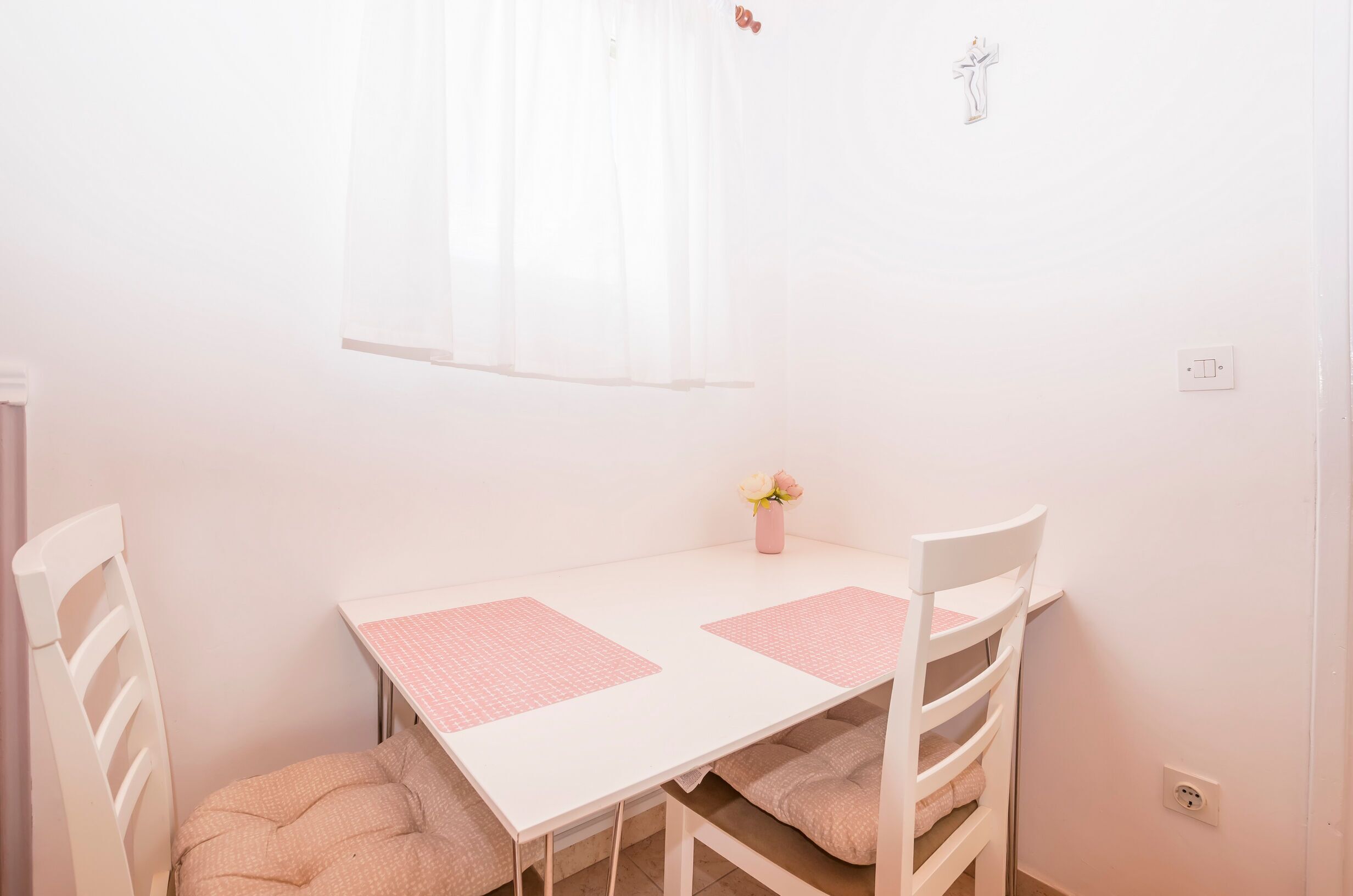 A small, white room with a table and two chairs. There is a window with sheer curtains, a small vase with flowers, and a wall-mounted light switch.